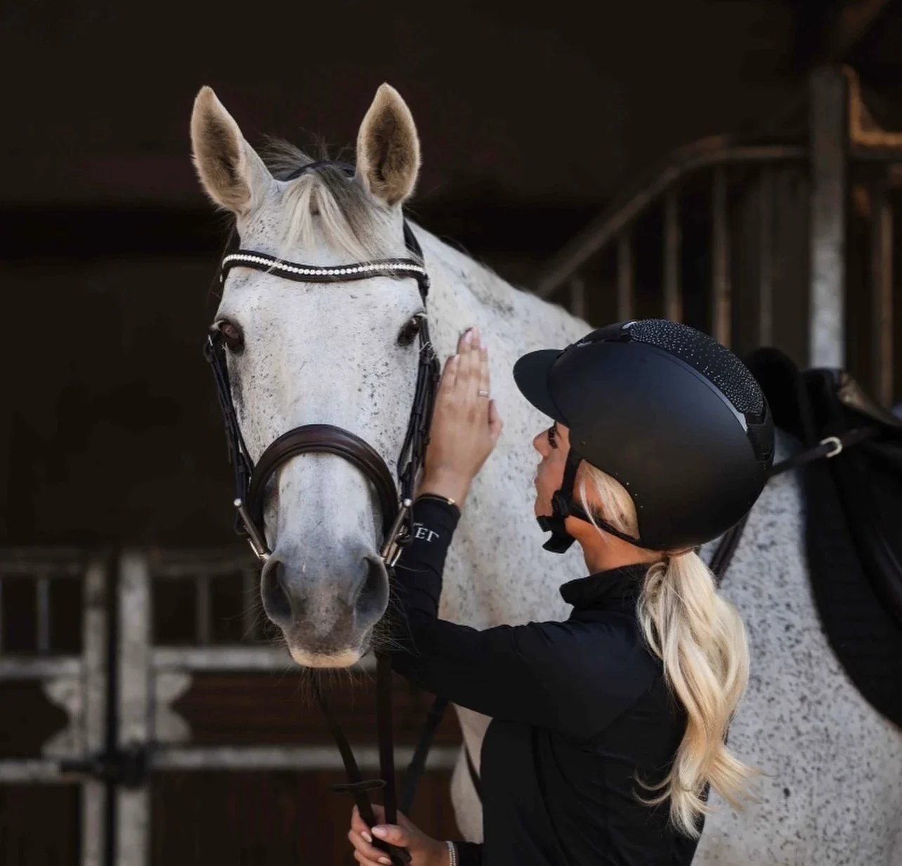 Ellie in a riding helmet gently petting a dapple-grey horse outside a stable